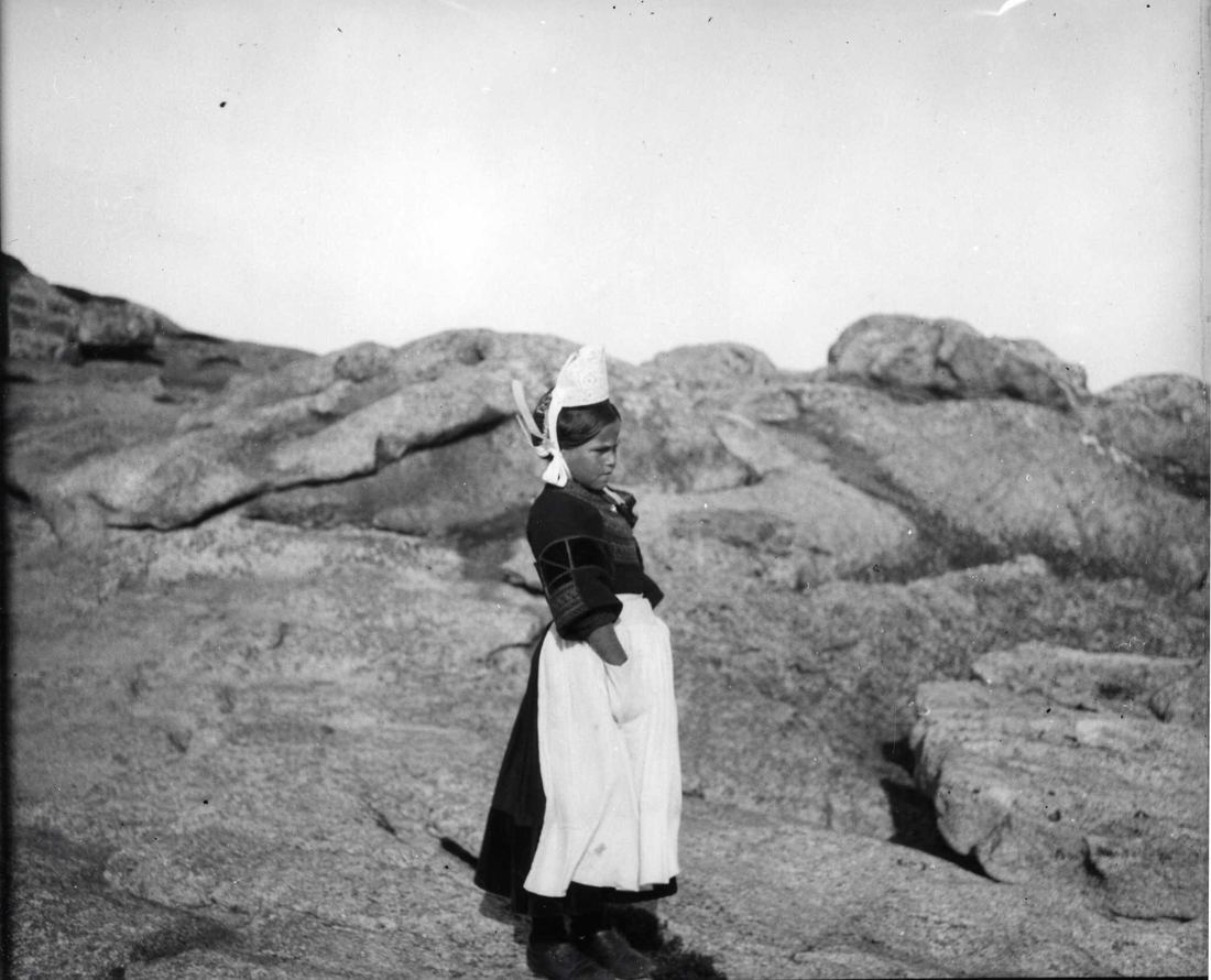 Petite Bigoudène sur les rochers de Saint-Guénolé, les mains dans le tablier — la coiffe est encore basse, celle d'avant les années 1920. La France est en guerre ; ici, le granit et le vent.